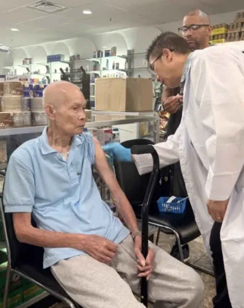 Pharmacist in a white coat preparing a vaccination for a seated older patient inside Patient Care Pharmacy